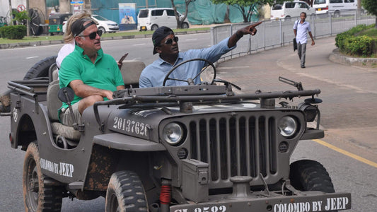 Two men in a vintage military-style jeep on a street with buildings and vehicles in the background.