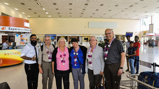 A group of five people standing in an airport terminal, likely at the arrival gate, with a man in a black suit and tie leading the group. Everyone is smiling and looking towards the camera.