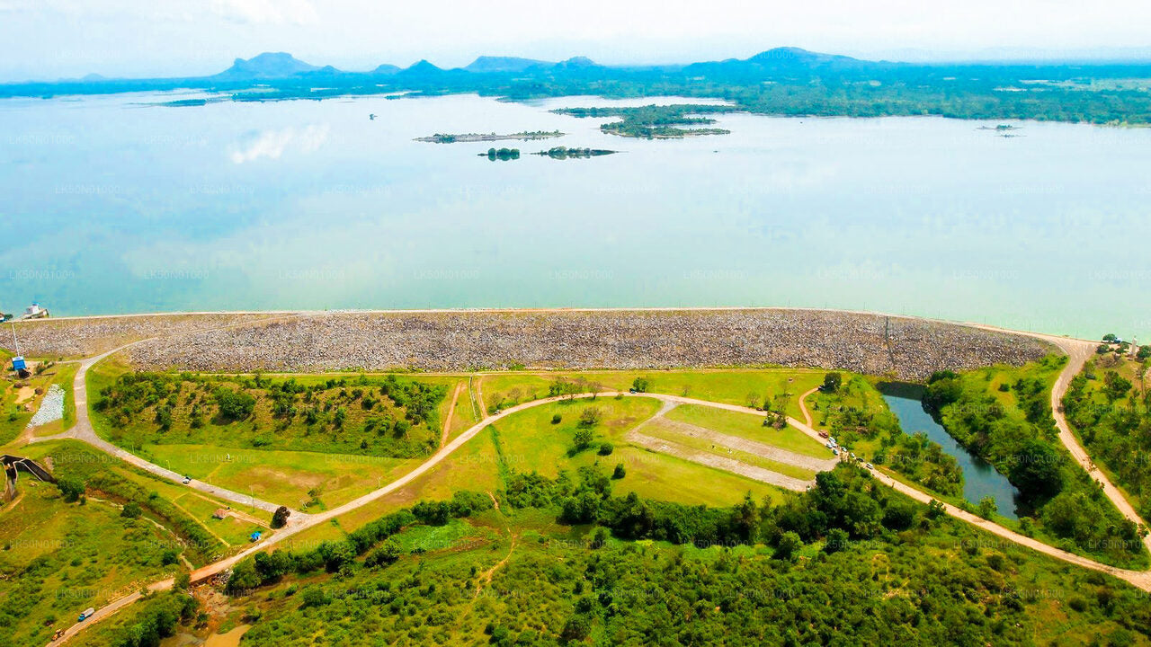 ALT text: Aerial view of a large reservoir with a long dam wall, surrounded by lush greenery, small roads, and distant mountains under a clear sky.
